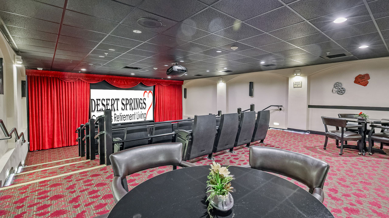 A small theater room with rows of black seats facing a stage with red curtains and a "Desert Springs Retirement Living" banner, carpeted floor, and round tables with plants in the foreground.