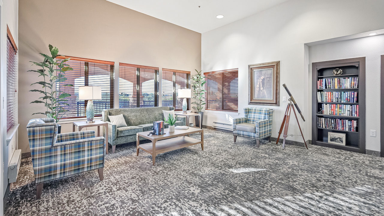 A bright, modern lounge with a sofa, two plaid armchairs, coffee tables, large windows with blinds, potted plants, bookshelves, a telescope on a stand, and framed art on the wall. Sunlight streams in.