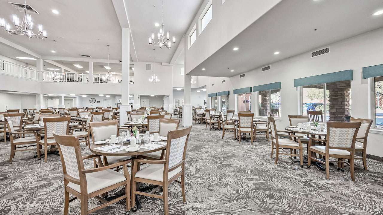 Spacious, well-lit dining room with multiple round tables and wooden chairs, neutral decor, large windows, chandeliers, and elegant place settings, creating a welcoming and modern atmosphere.