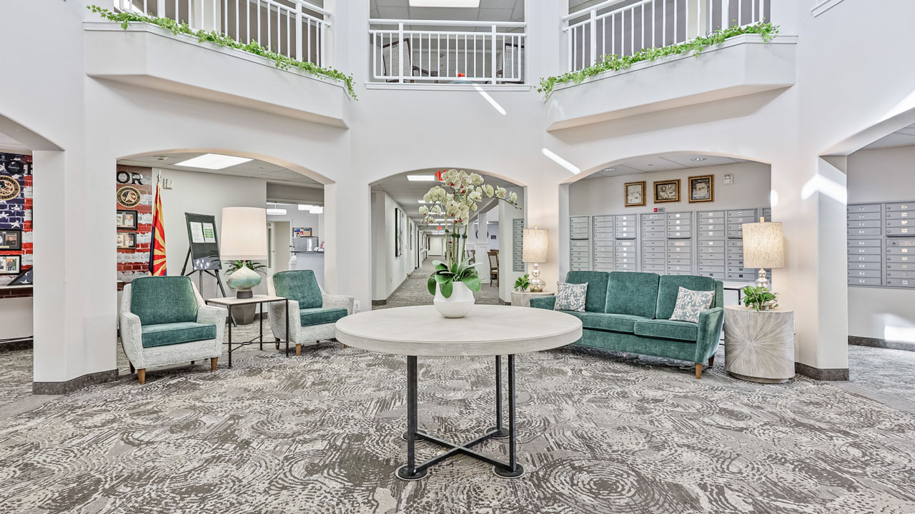Bright, spacious lobby with a round table and floral centerpiece, green sofas and chairs, lamps, mailboxes, decorative plants on balconies, and patterned carpet. White walls and high ceiling create an airy, welcoming atmosphere.