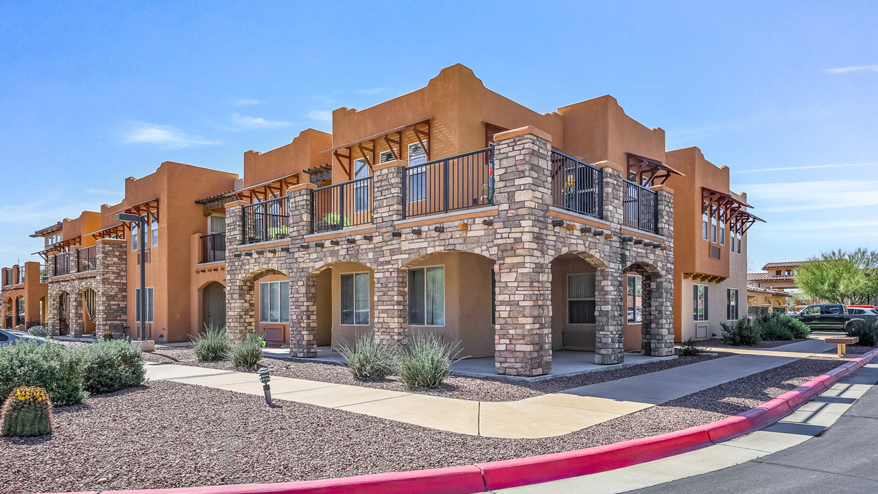 A modern southwestern-style apartment building with stone arches, stucco walls, balconies, and desert landscaping under a clear blue sky.