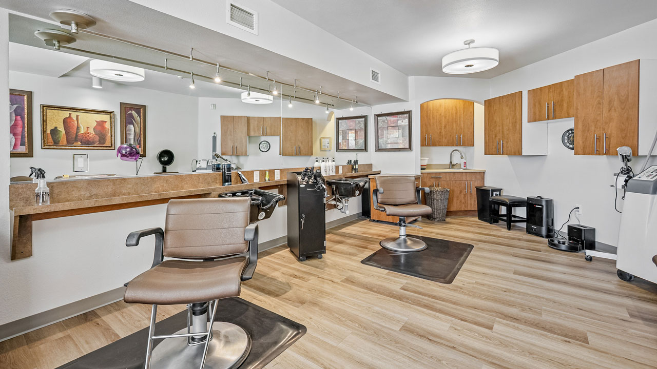 Modern hair salon with two brown styling chairs, wooden floors, a large mirror, wooden cabinets, framed art on the walls, and bright overhead lighting. The space is clean, organized, and inviting.