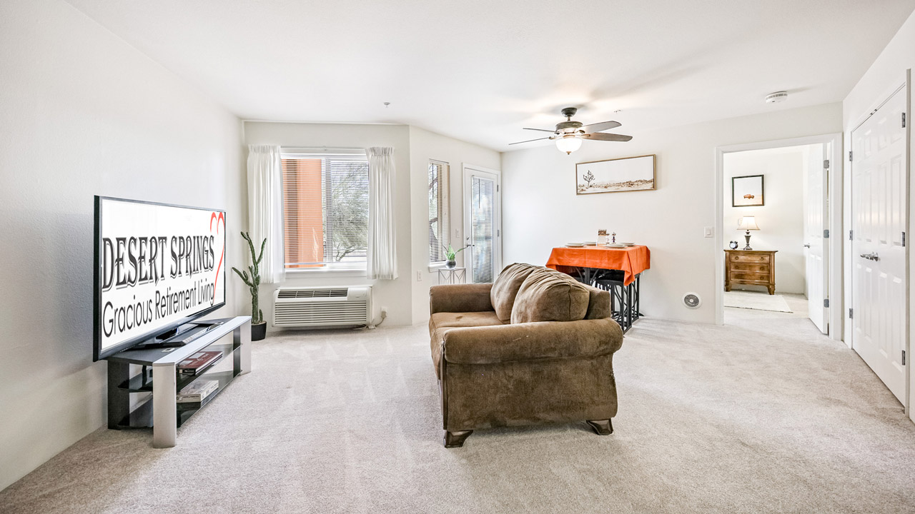 A bright, carpeted living room with a brown sofa facing a TV displaying "Desert Springs Gracious Retirement Living." Large windows, a ceiling fan, a potted cactus, and a doorway to a bedroom are visible.