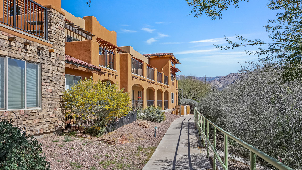 A paved walkway with a green railing runs alongside a southwestern-style building with earth-toned walls, stone accents, and balconies, surrounded by shrubs and desert plants under a clear blue sky.