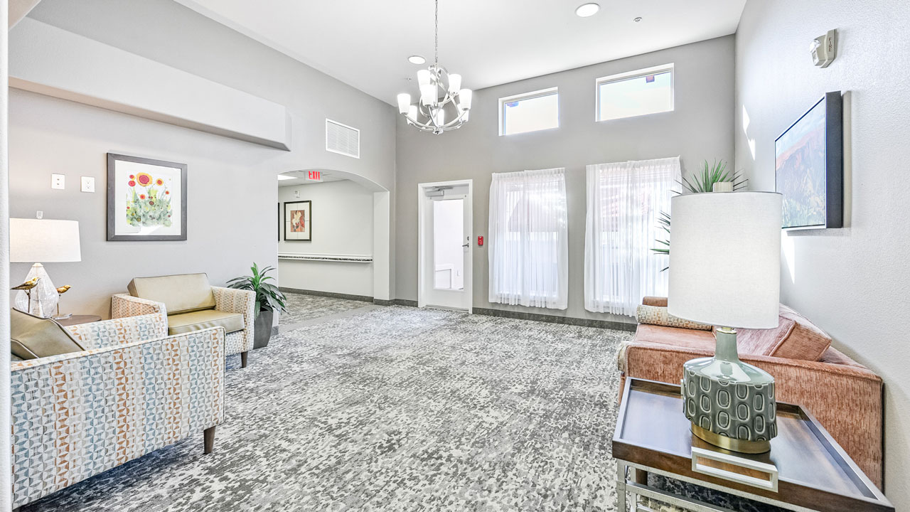 A bright, modern lounge area with armchairs, a sofa, decorative lamps, wall art, plants, and large windows with sheer curtains letting in natural light. A chandelier hangs from the ceiling.