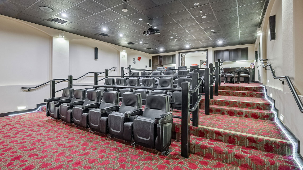 A modern, small movie theater with rows of black cushioned seats, red patterned carpet, and lighting along the steps. The room has a projector mounted on the ceiling and a small kitchenette area at the back.