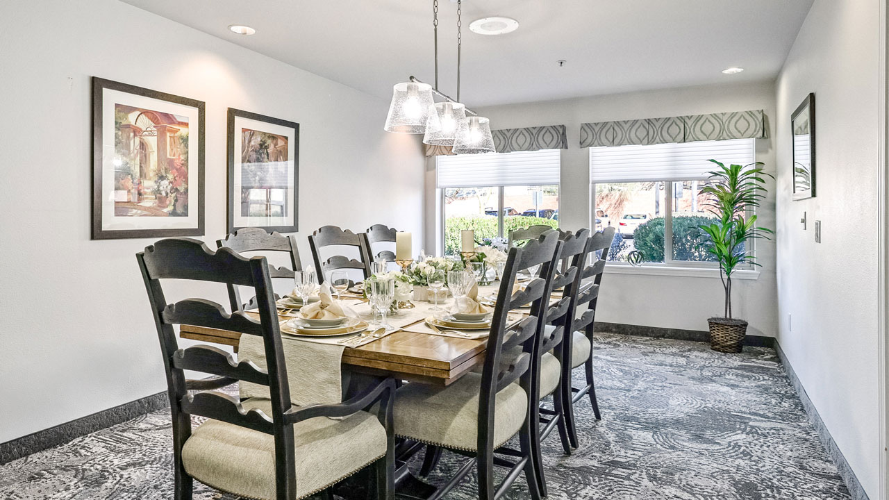 A bright dining room with a long wooden table set for eight, cushioned chairs, decorative tableware, artwork on the walls, large windows letting in sunlight, and a potted plant in the corner.