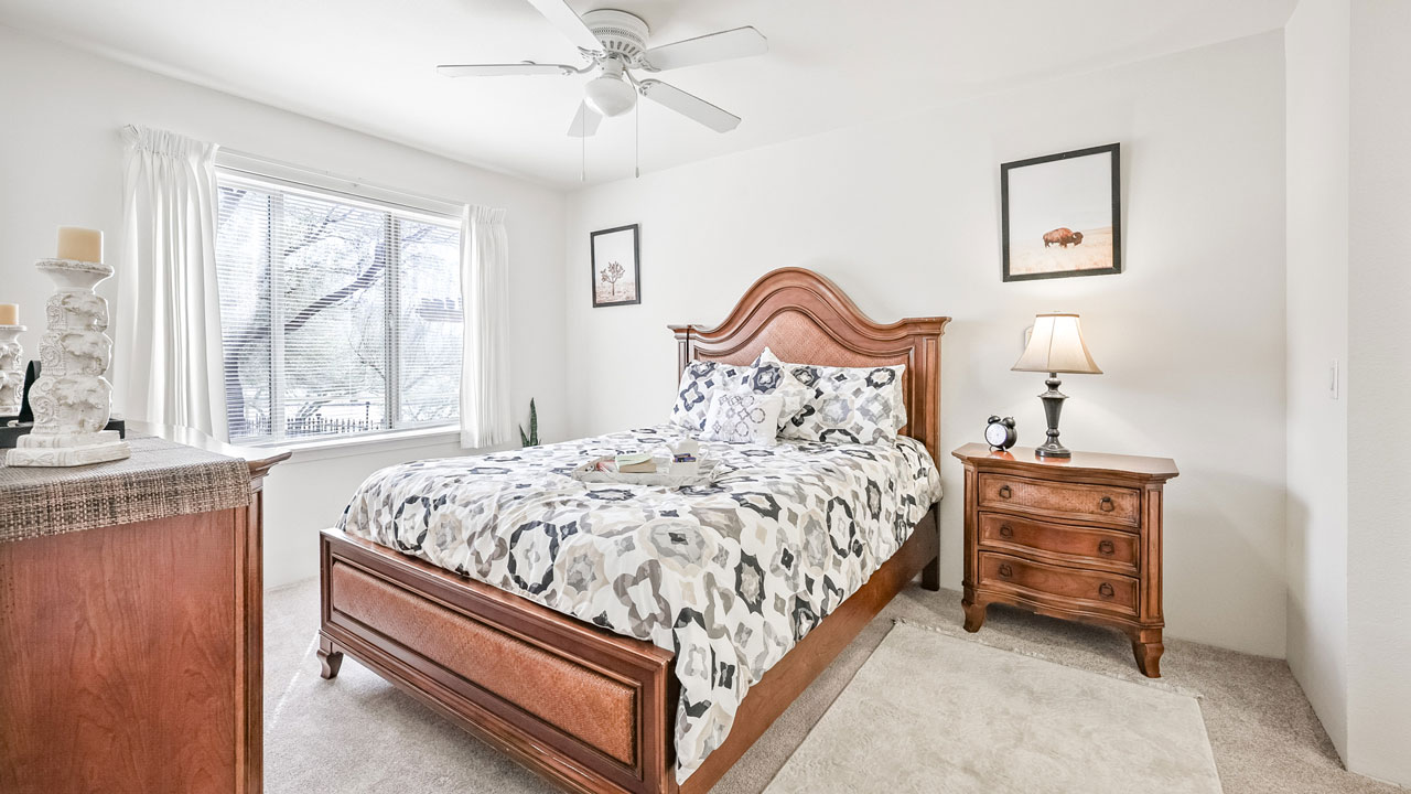 A bright bedroom with a wooden bed frame, patterned bedding, a matching nightstand with a lamp, a dresser, a ceiling fan, light carpet, and a large window with white curtains letting in natural light.