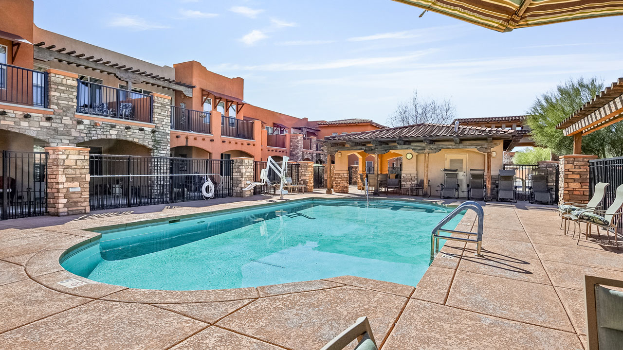 Outdoor swimming pool surrounded by a stone patio, lounge chairs, and a safety fence, with a multi-story building featuring balconies in the background under a clear blue sky.