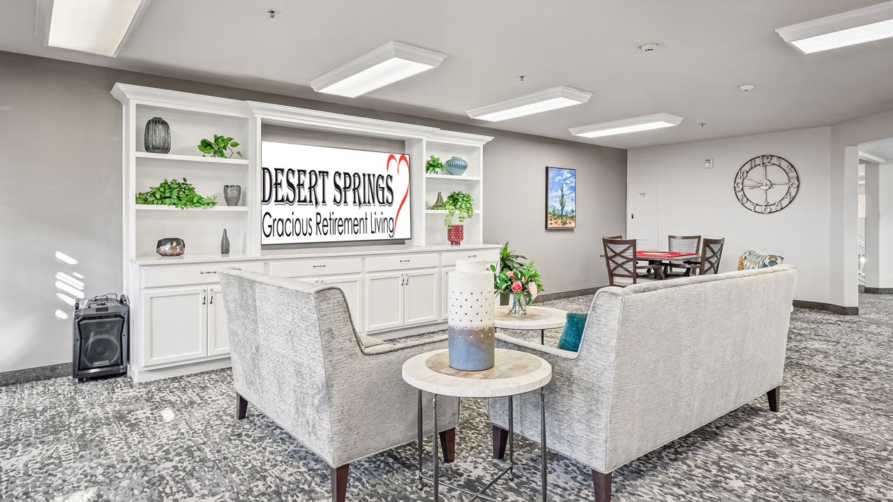 A bright, modern lounge with grey sofas, a white built-in shelving unit displaying “DESERT SPRINGS Gracious Retirement Living,” a round table with chairs, plants, and a decorative wall clock.