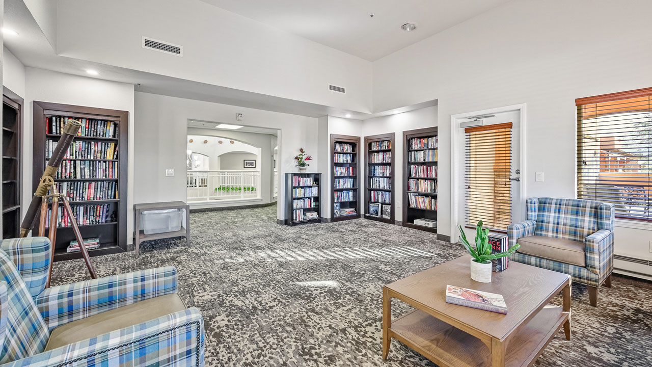 A bright library room with four tall bookshelves, two plaid armchairs, a wooden coffee table with a plant, a telescope, and large windows allowing natural light in. An open doorway leads to a hallway outside.