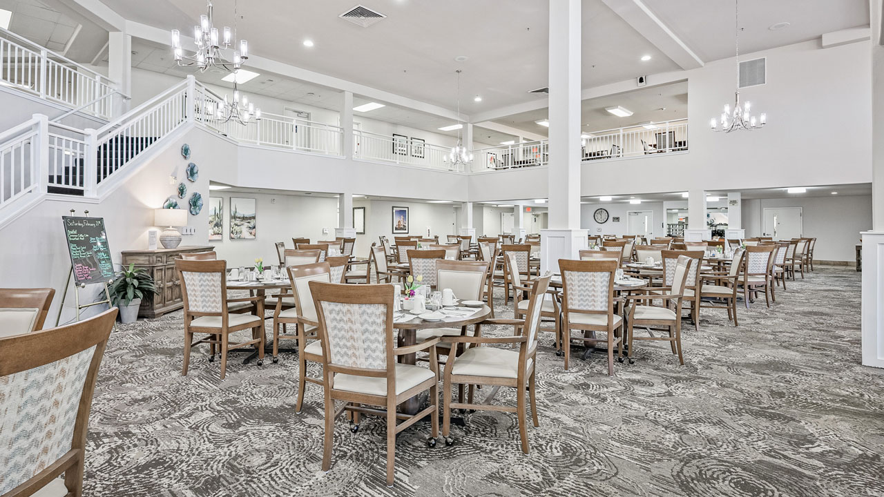 Spacious, elegant dining room with round tables and wooden chairs. Light walls, chandeliers, patterned carpet, and a staircase leading to an upper mezzanine. The tables are set for dining with cups, plates, and flowers.