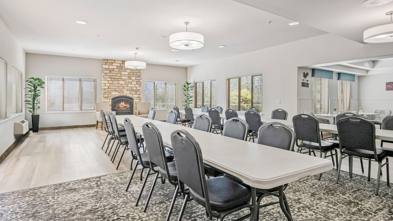 A bright dining room with long white tables, black chairs, large windows, a stone fireplace, two tall plants, and modern ceiling lights. The space has light wood flooring and a patterned rug.