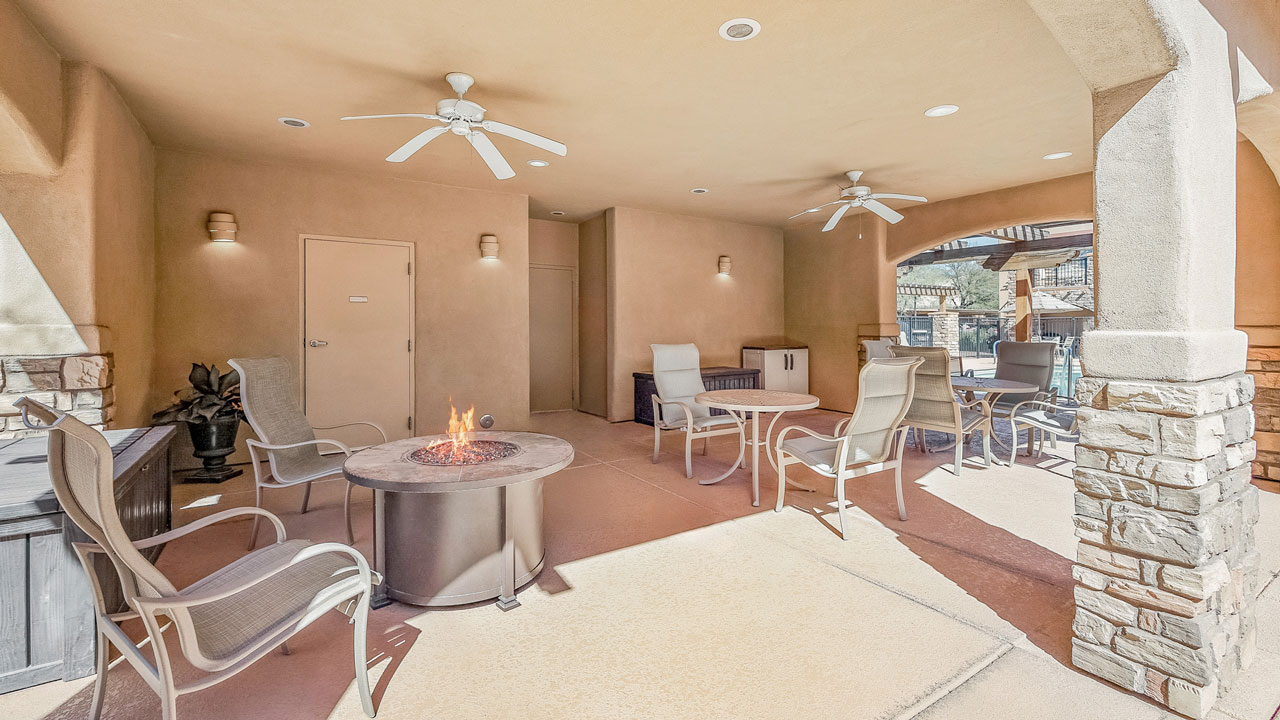 A covered outdoor patio area with beige walls, ceiling fans, and several chairs arranged around a round fire pit table. Sunlight filters in, and stone columns frame the open space.