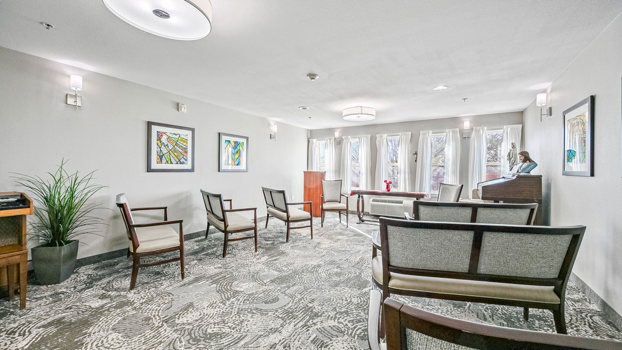 A bright, modern room with several chairs arranged in rows, framed artwork on the walls, large windows with sheer curtains, and a woman sitting on a piano bench in the corner. A potted plant and wooden cabinet are also visible.
