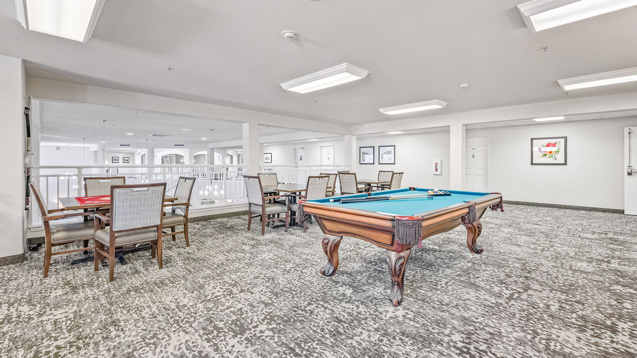 A spacious, carpeted game room with a pool table in the center and several tables with chairs along the side. The room has bright lighting and framed pictures on white walls.