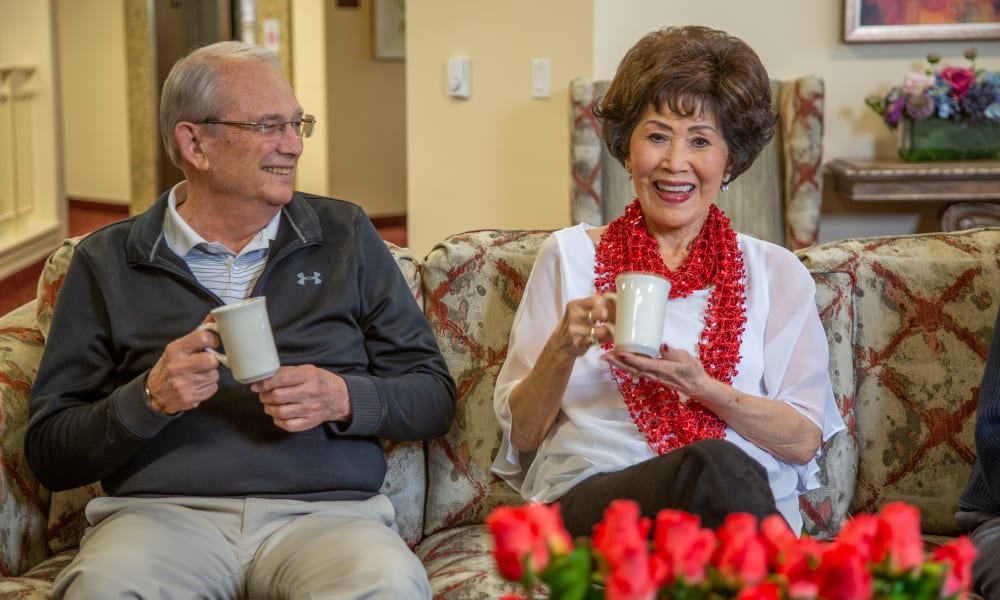 An older man and woman are sitting on a floral-patterned sofa, smiling and holding white mugs. The woman is wearing a red scarf and a white blouse, while the man is dressed in a grey sweater. There are red flowers in the foreground and a cozy living room in the background.