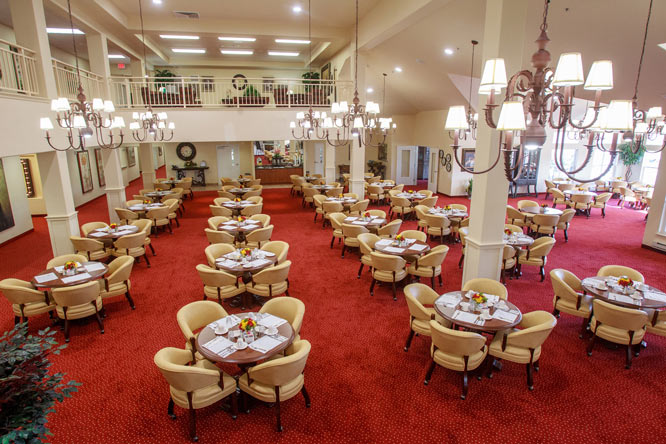 A large dining hall with red carpet features numerous round tables set for four. Each table is equipped with beige chairs and is adorned with small floral centerpieces. Chandeliers hang from the high ceiling, and a balcony overlooks the space.