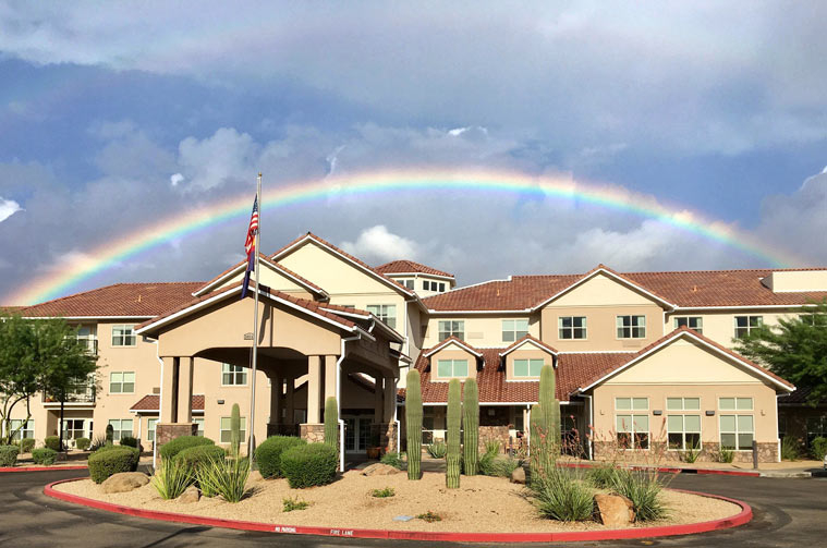 A modern, multi-story building with a beige facade, visible through a landscaped foreground with desert plants like cacti. A rainbow arches across the cloudy sky above the building. An American flag is flying on a flagpole near the entrance.