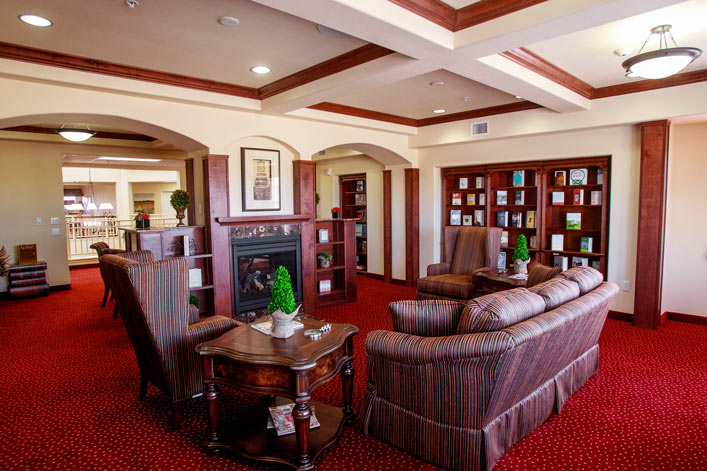 A cozy living area featuring striped upholstered couches and chairs around a wooden coffee table and fireplace. Bookshelves line the walls, and a soft red carpet covers the floor. Potted plants and framed artwork add a homey touch under warm ceiling lights.