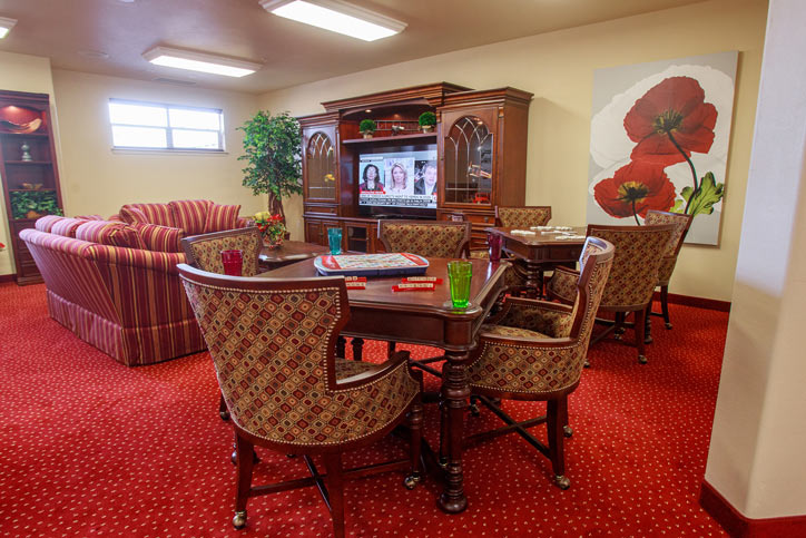 A cozy living room features a wooden entertainment center with a television, floral artwork, red sofas, a game table with board games, and green plants. The red carpet and soft lighting create a warm and inviting atmosphere.