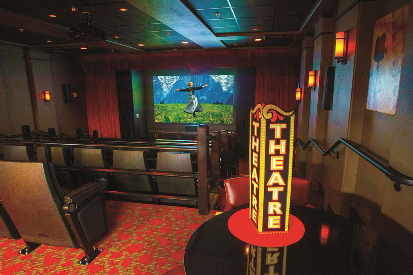 A home theater room with theater seating, red carpet with a patterned design, and red curtains. A brightly lit marquee sign with the word 