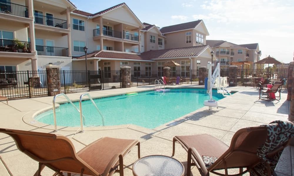 Outdoor swimming pool with lounge chairs and tables around it at an apartment complex. The apartment buildings have balconies and beige-colored walls with red-tiled roofs. A safety rail and pool lift are present next to the pool. The sky is partly cloudy.