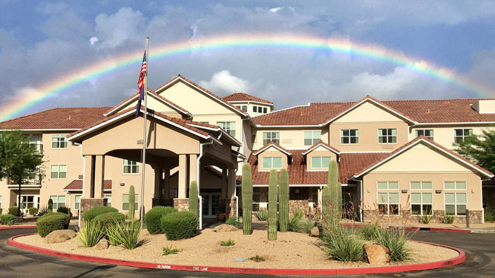 A three-story building with a reddish-brown roof and a front entrance canopy is set against a partly cloudy sky with a vivid rainbow arching across. The landscape features desert vegetation, including cacti and shrubs, with an American flag flying near the entrance.