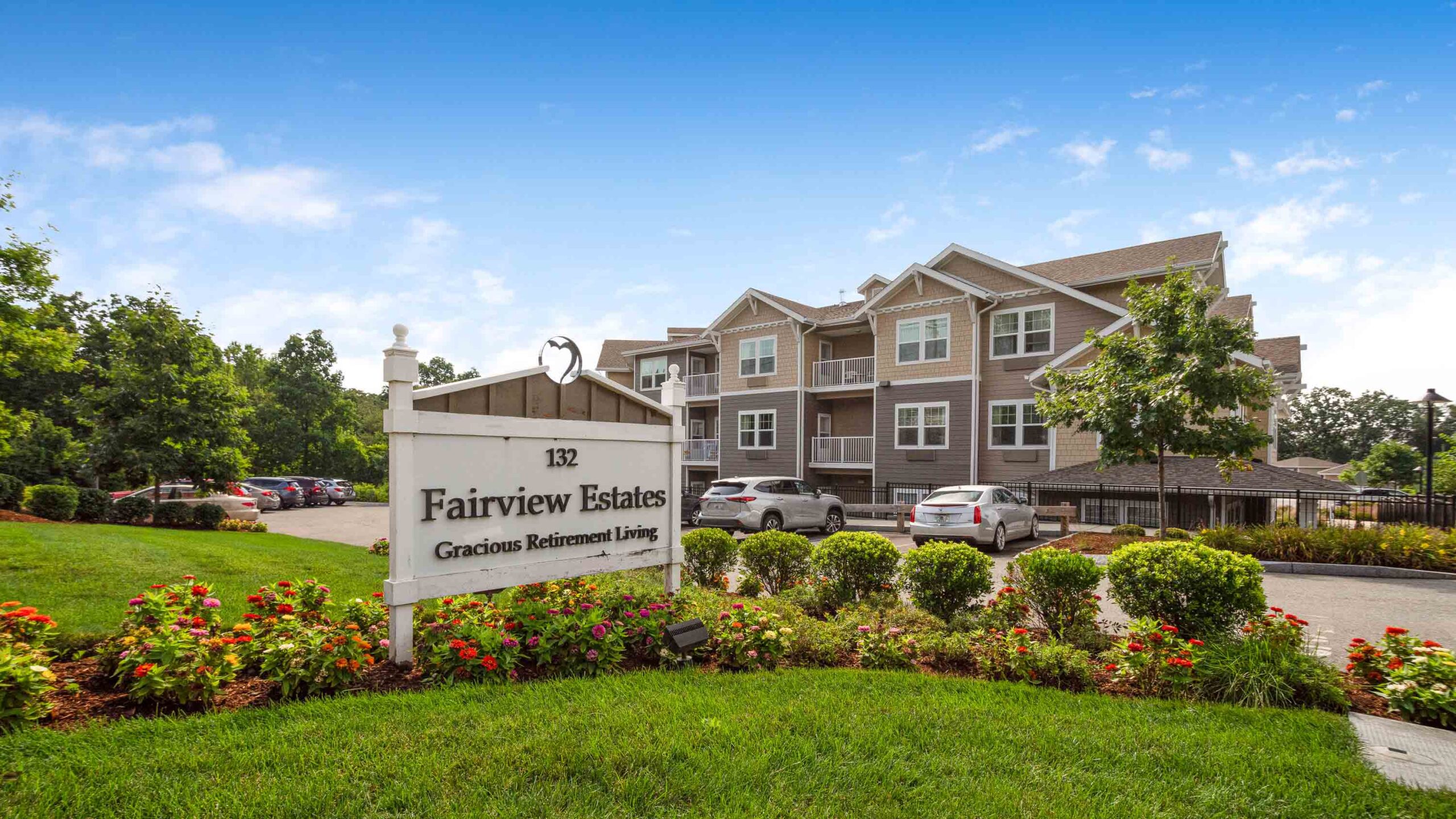 A sign reading "Fairview Estates Gracious Retirement Living" stands in a landscaped garden, with a multi-story residential building and parked cars in the background under a blue sky.