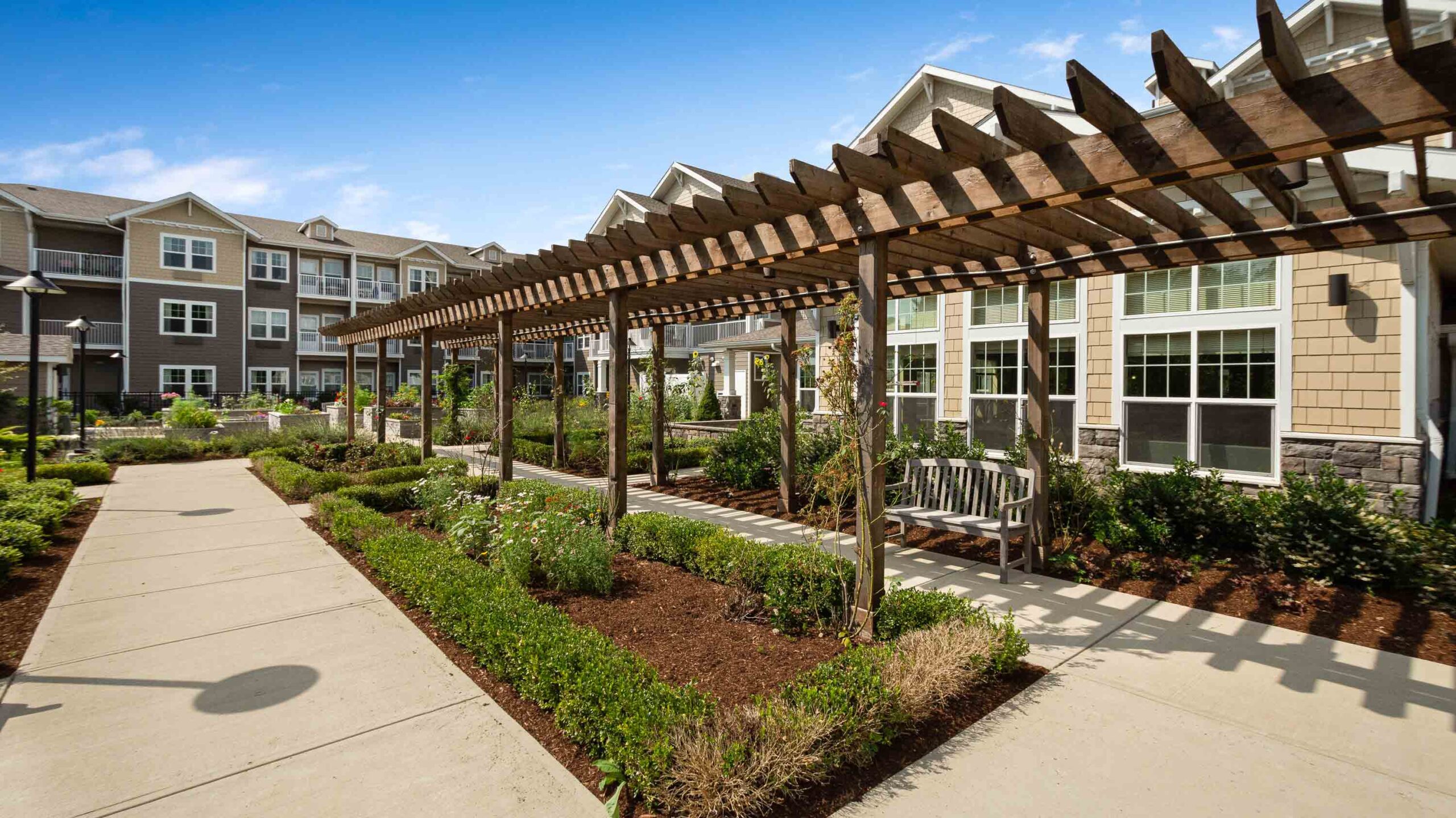 A landscaped courtyard with a wooden pergola, benches, and neatly trimmed bushes, next to a modern apartment building under a blue sky.
