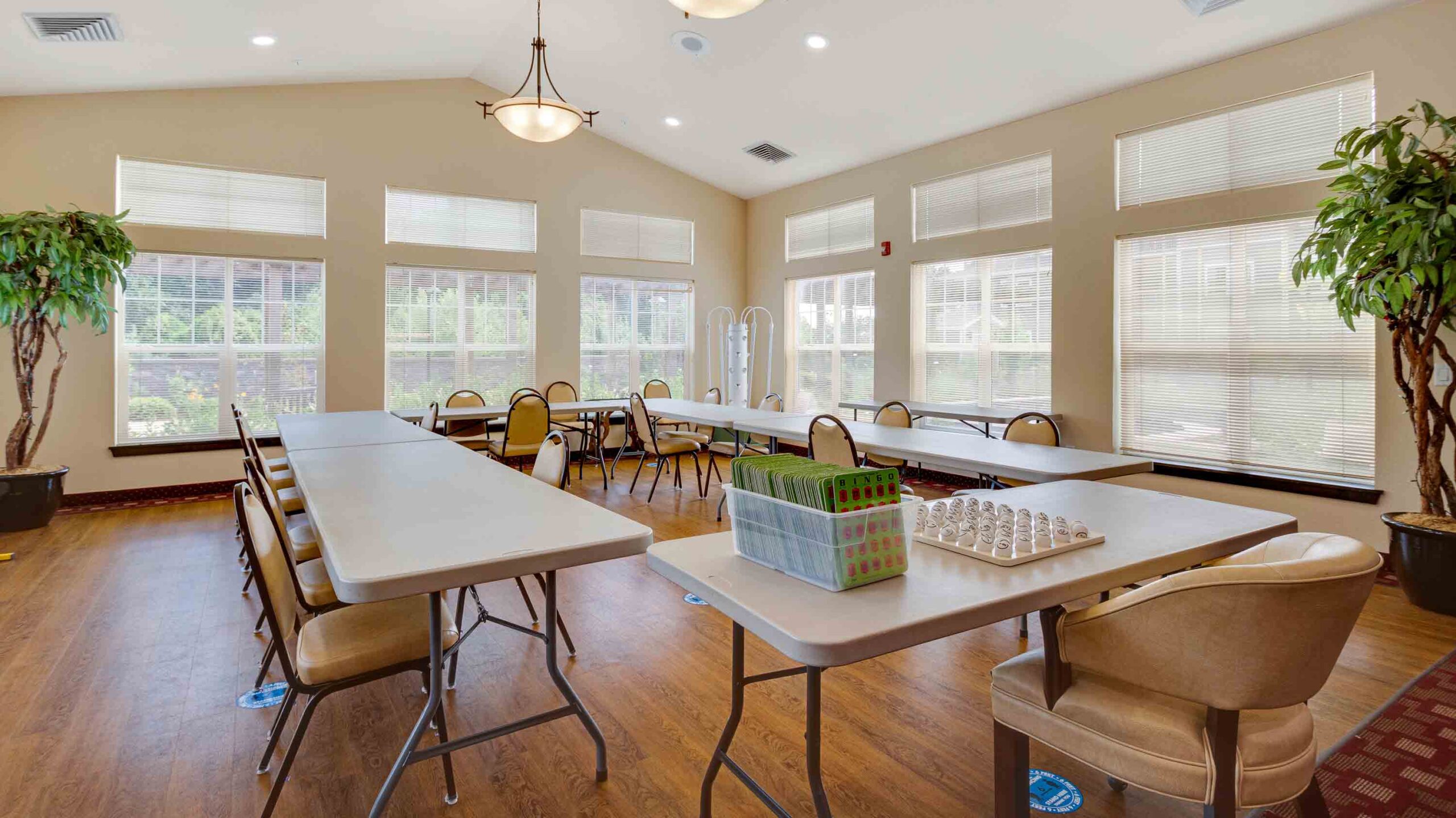 A bright, spacious room with large windows, multiple rectangular tables and chairs, chess and Connect Four games set up, wooden floor, green potted plants, and overhead lighting.