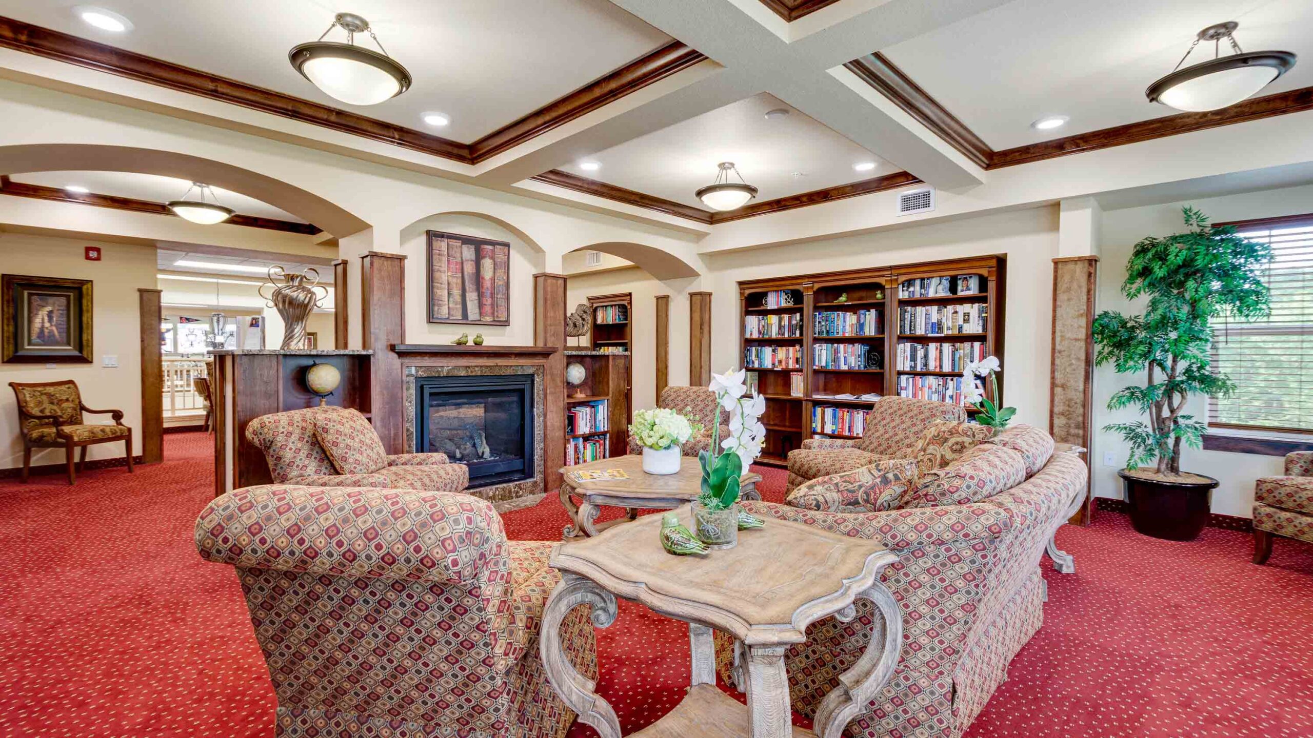 A cozy library lounge with patterned armchairs around a wooden table, bookshelves along the walls, a fireplace, red carpet, indoor plants, and warm lighting from ceiling fixtures.