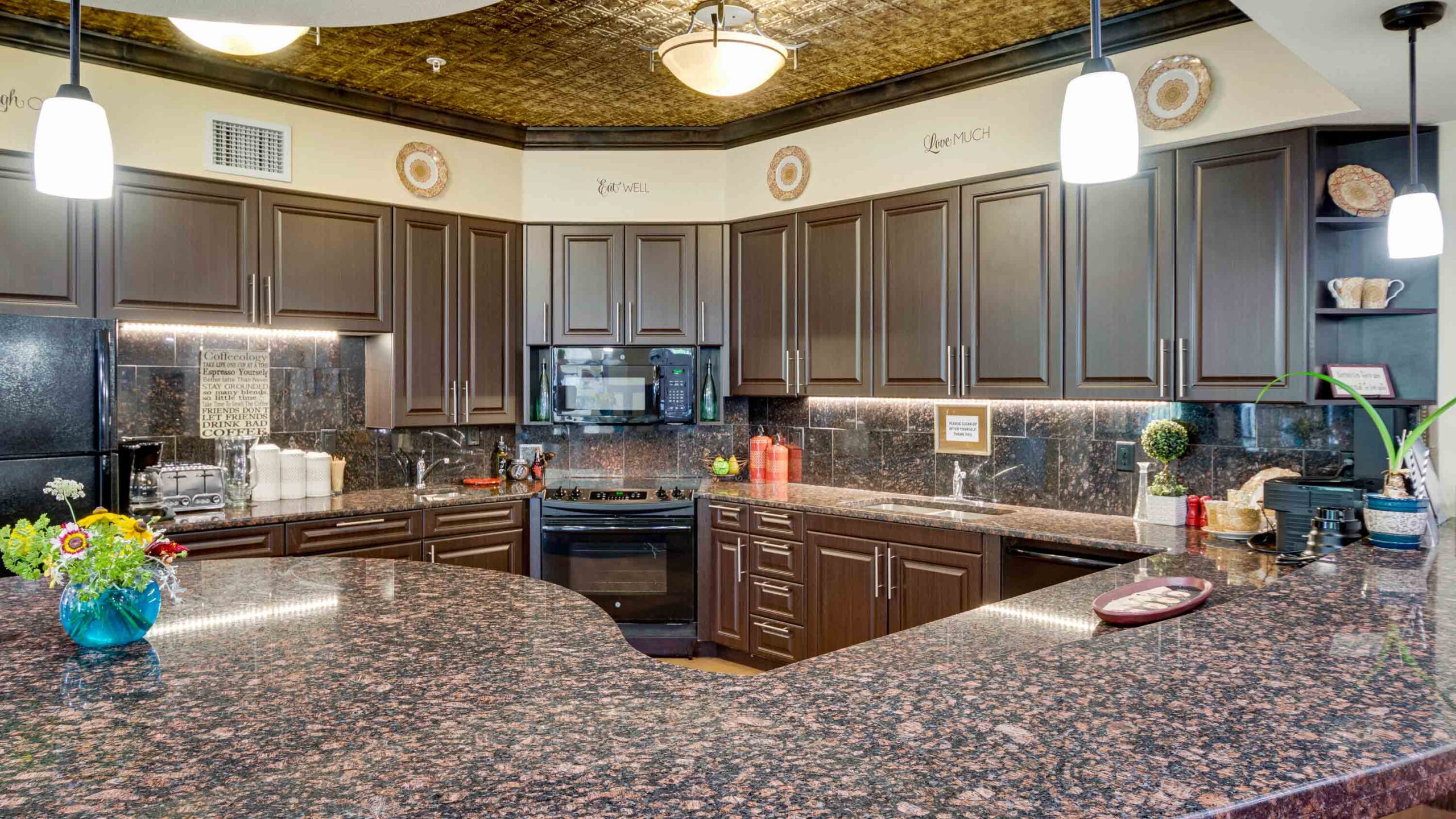 Spacious kitchen with dark wood cabinets, black appliances, granite countertops, pendant lights, decorative plates above cabinets, and a vase of flowers on the counter.