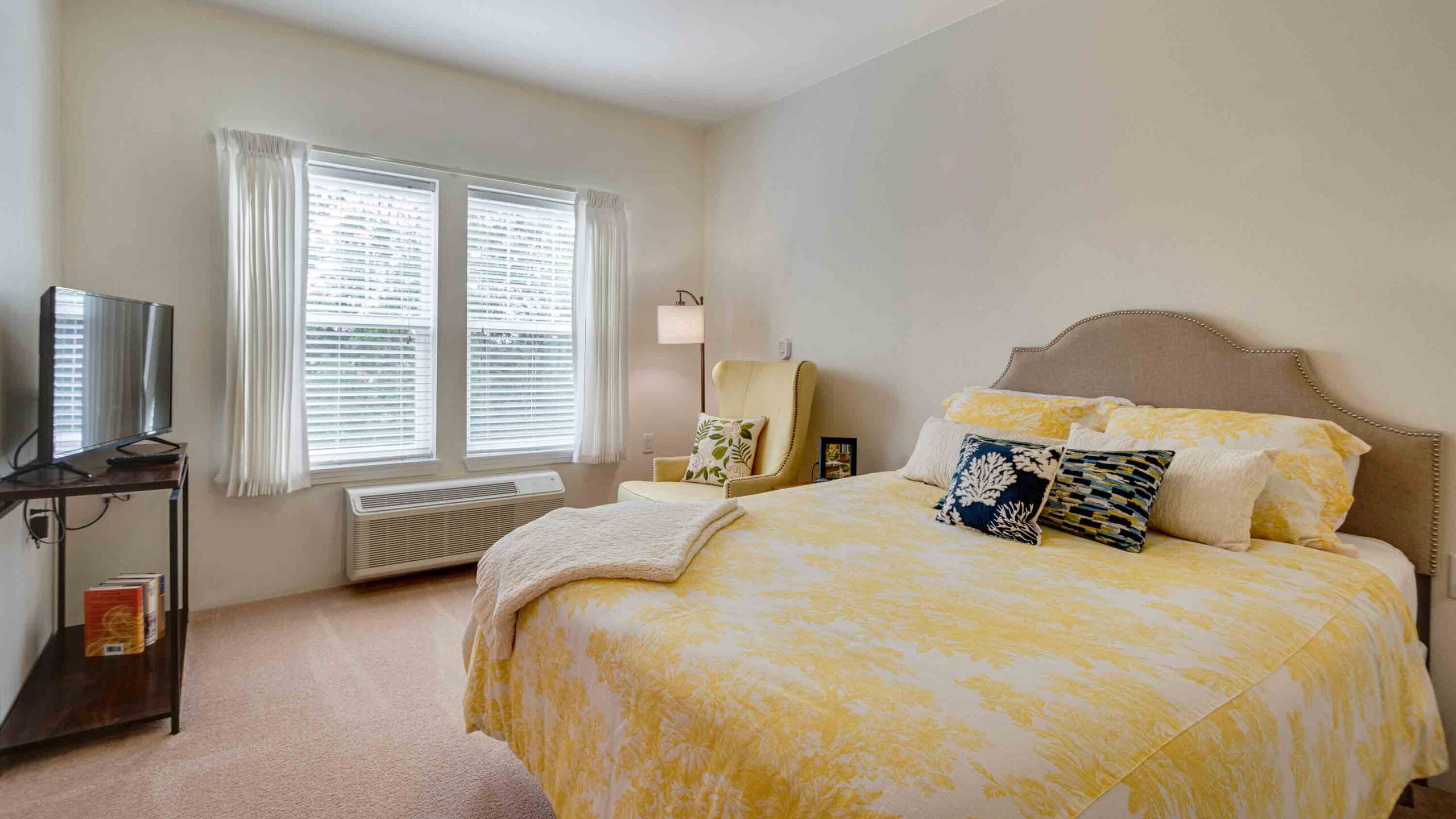 A cozy bedroom with a bed covered in yellow and white bedding, decorative pillows, a TV on a stand, an armchair with a cushion, a floor lamp, and a large window with white curtains letting in natural light.