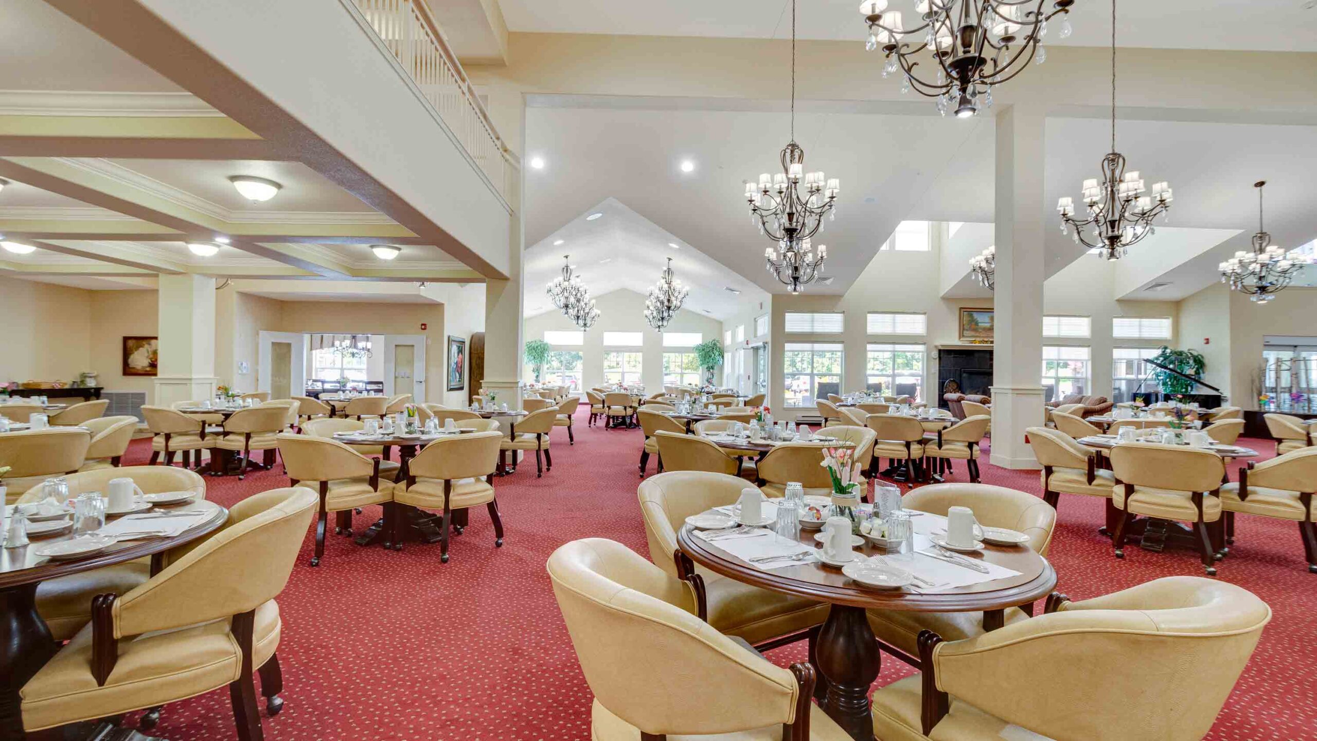 Spacious, well-lit dining room with round tables set for meals, beige upholstered chairs, red carpet, large windows, and elegant chandeliers hanging from high ceilings.