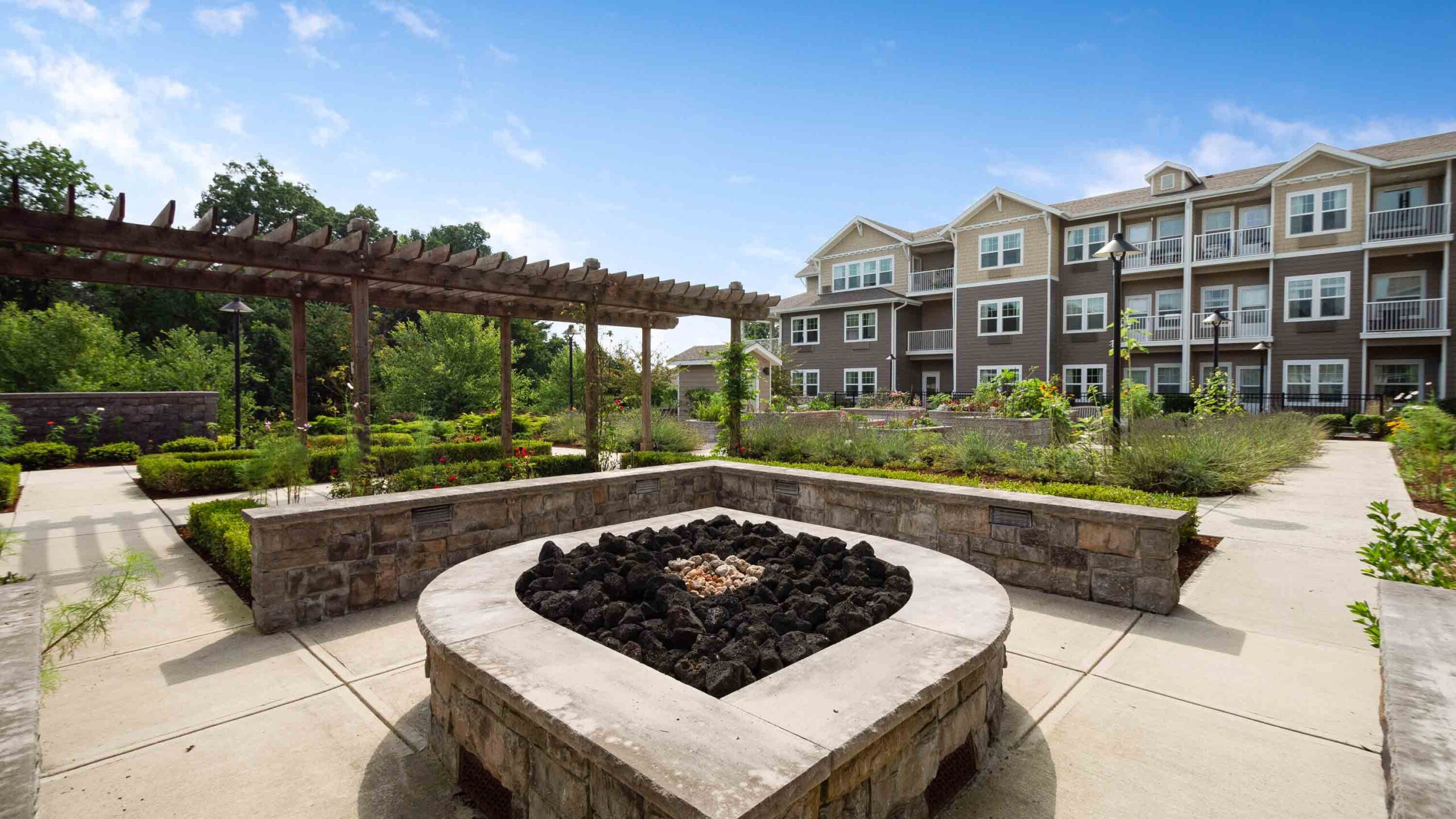 A landscaped courtyard with a stone fire pit, wooden pergola, and walkways, surrounded by greenery and a three-story apartment building in the background under a blue sky.