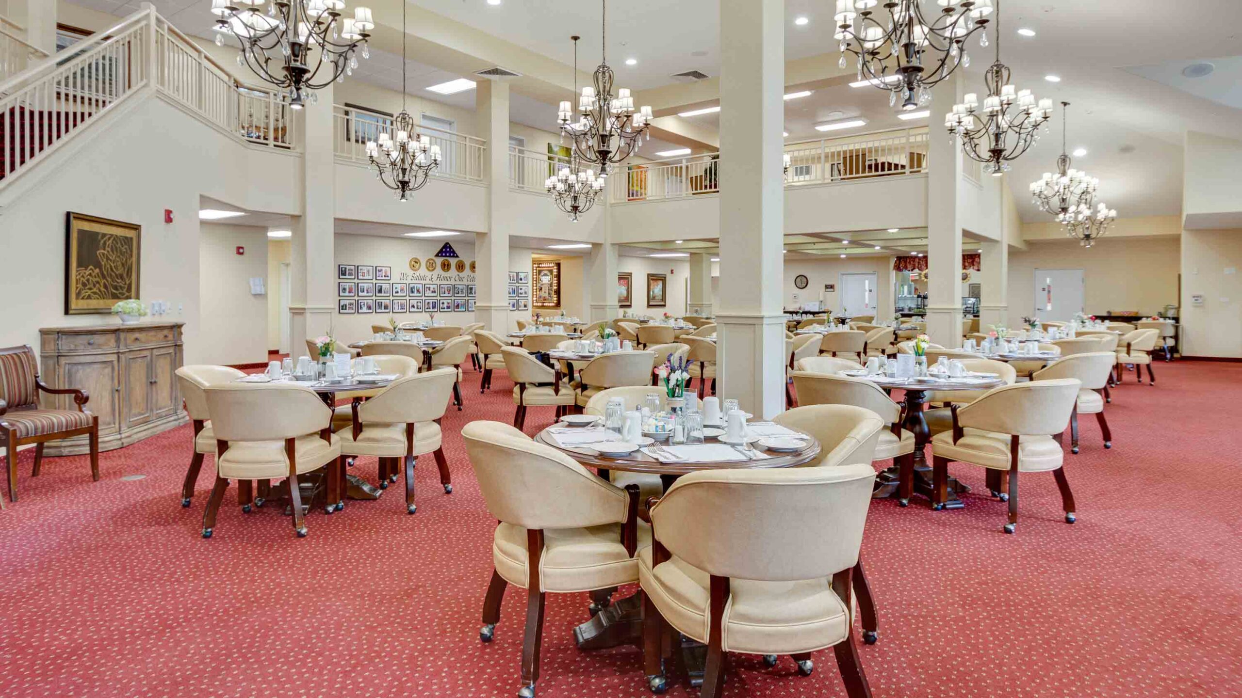 Spacious dining room with round tables set for meals, cream-colored chairs, red carpet, and chandeliers hanging from a high ceiling. A balcony and framed pictures are visible in the background.