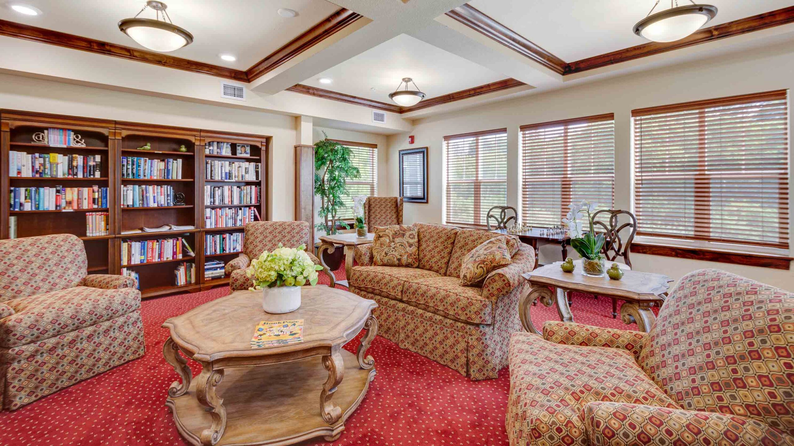 A cozy lounge with patterned armchairs and sofas, a wooden coffee table with a flower arrangement, bookshelves filled with books, large windows with blinds, and potted plants on red carpeted flooring.