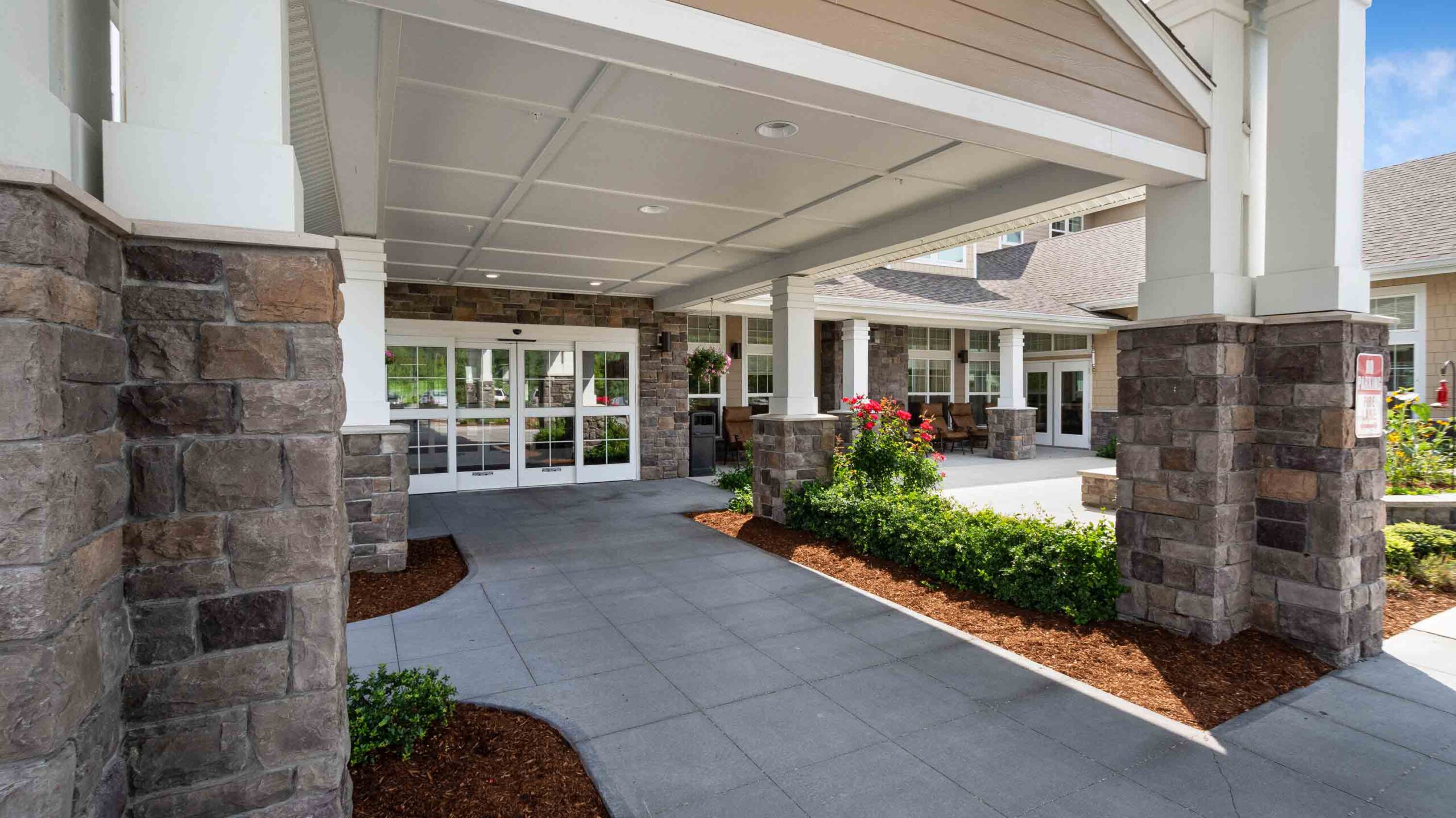 Covered entrance of a modern building with stone pillars, glass doors, and neatly landscaped flowerbeds along the walkway leading to the entrance.