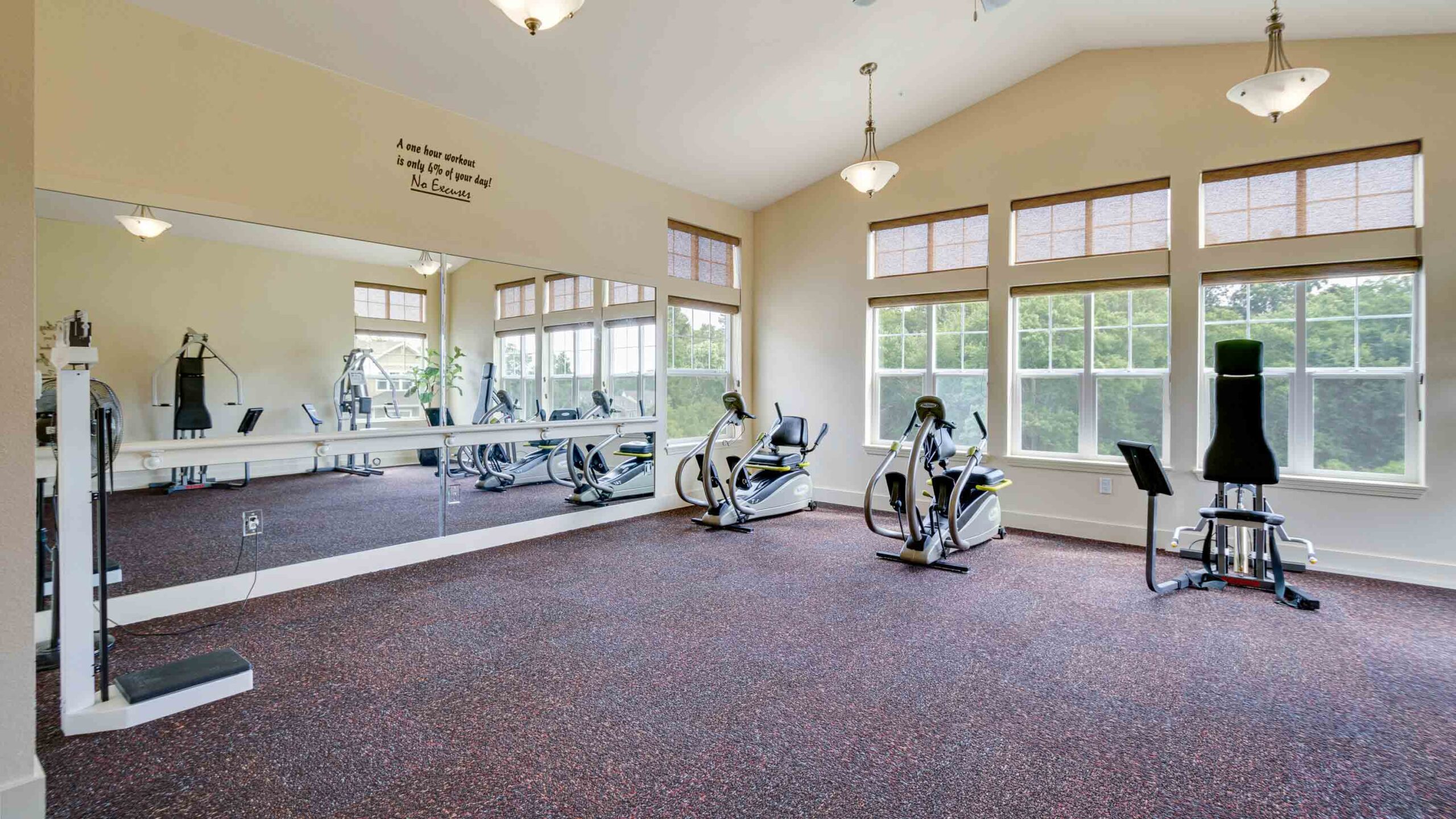 Spacious gym room with large mirrors, two exercise bikes, a weight machine, and big windows letting in natural light onto a speckled floor.