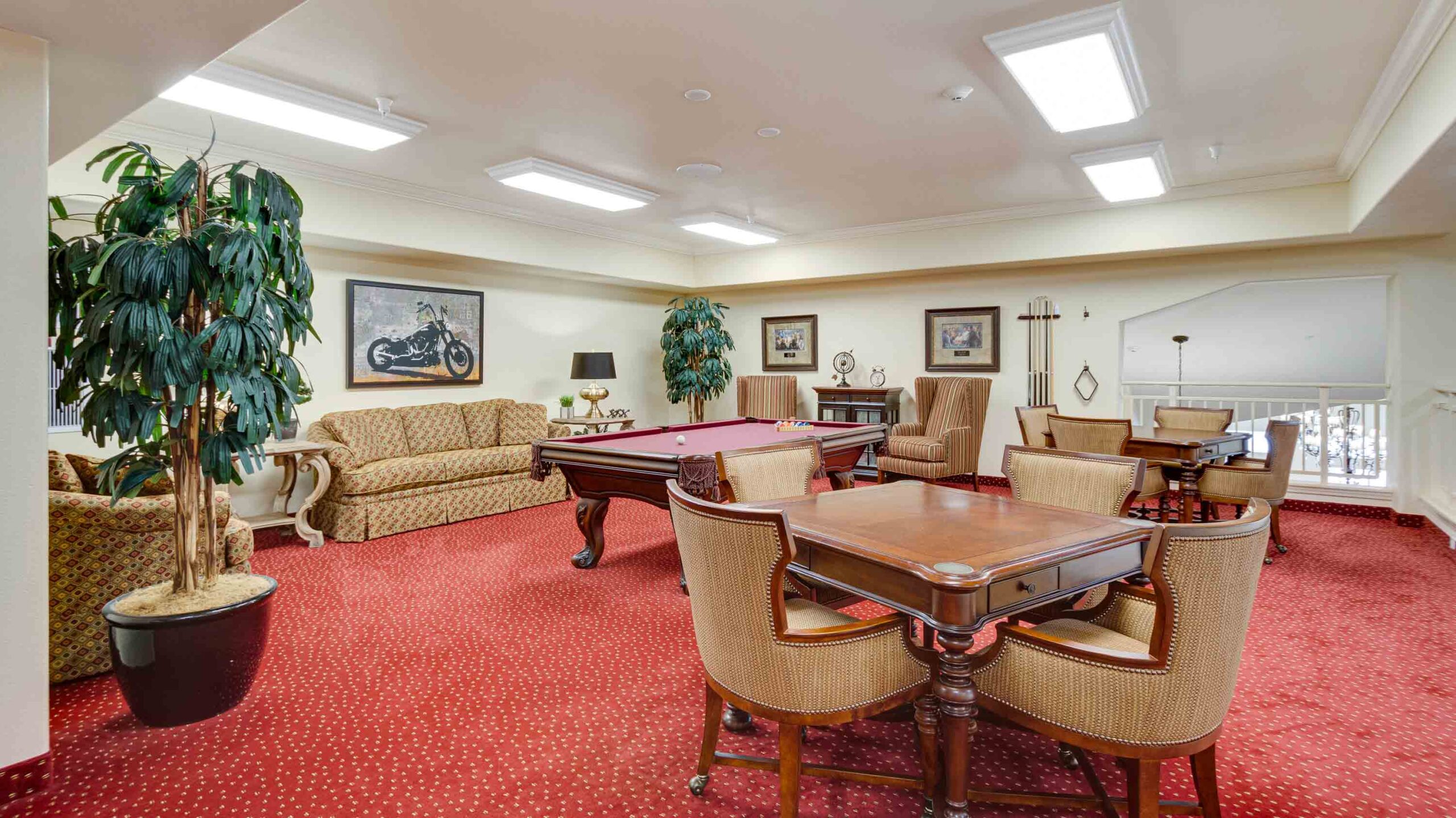 A spacious lounge with red carpet, a pool table, several wooden tables and chairs, a beige sofa, framed artwork on the walls, potted plants, and bright ceiling lights.