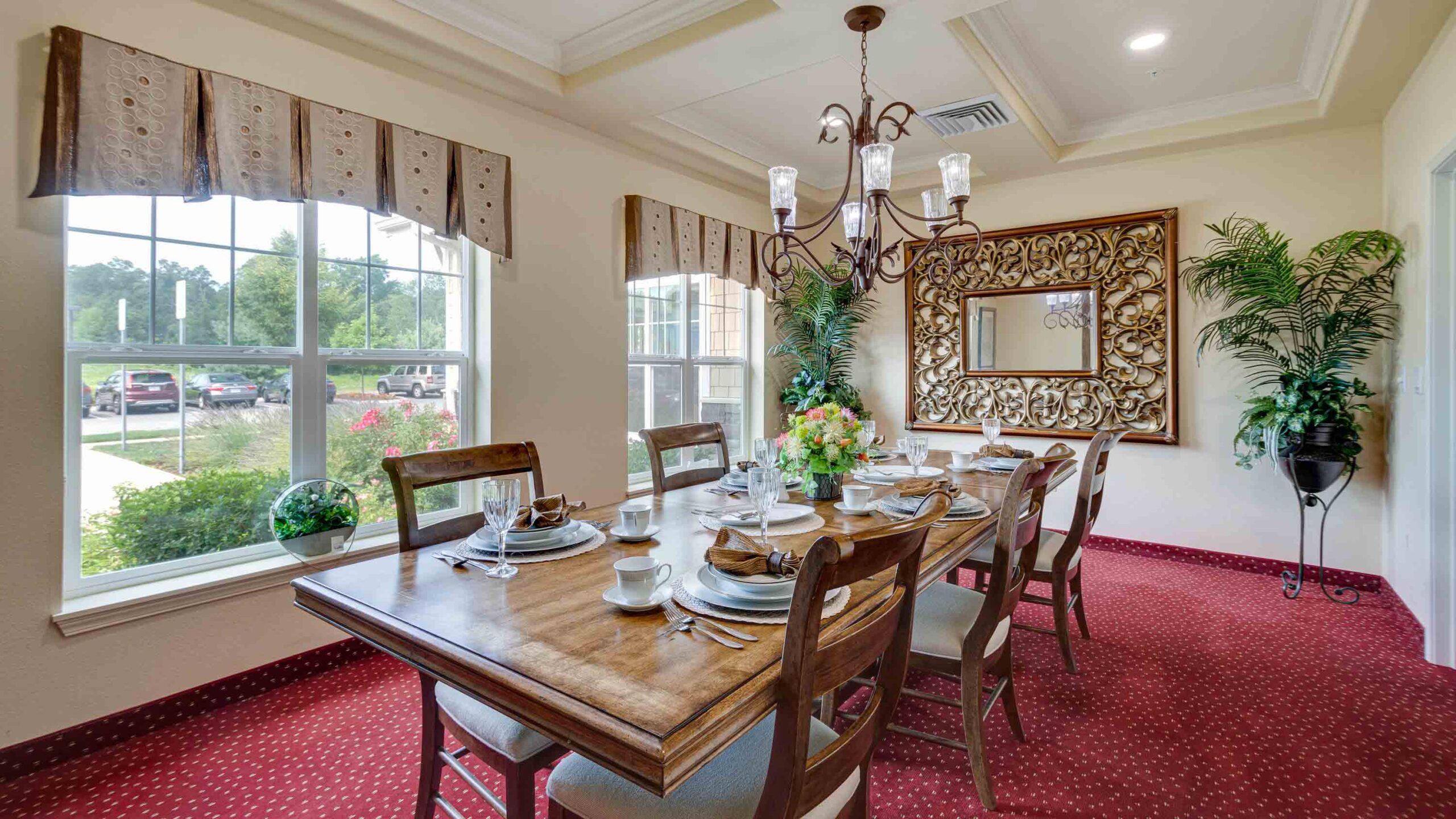 A dining room with a wooden table set for six, cushioned chairs, red carpet, large decorative mirror, green plants, chandelier, and windows letting in natural light.