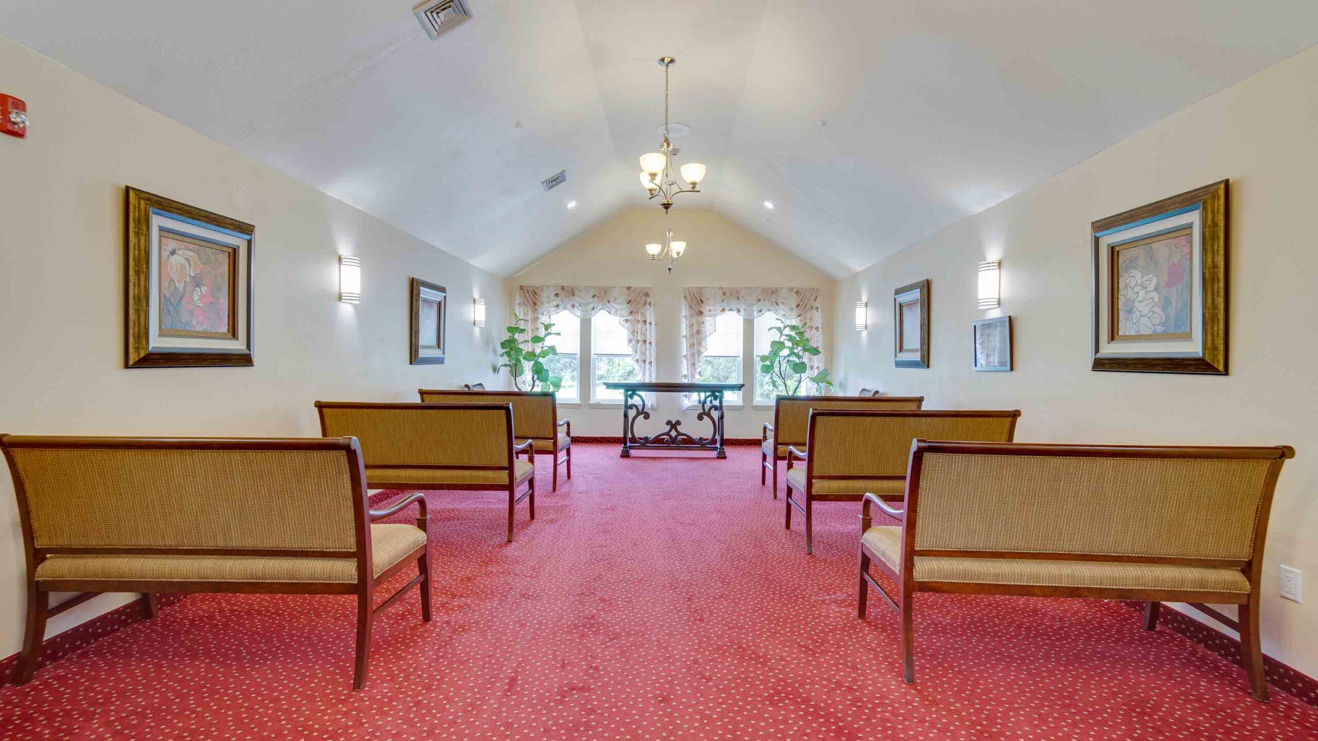 A bright, carpeted room with benches facing a table near large windows, decorated with framed art, plants, and chandeliers, arranged symmetrically for a formal or gathering space.