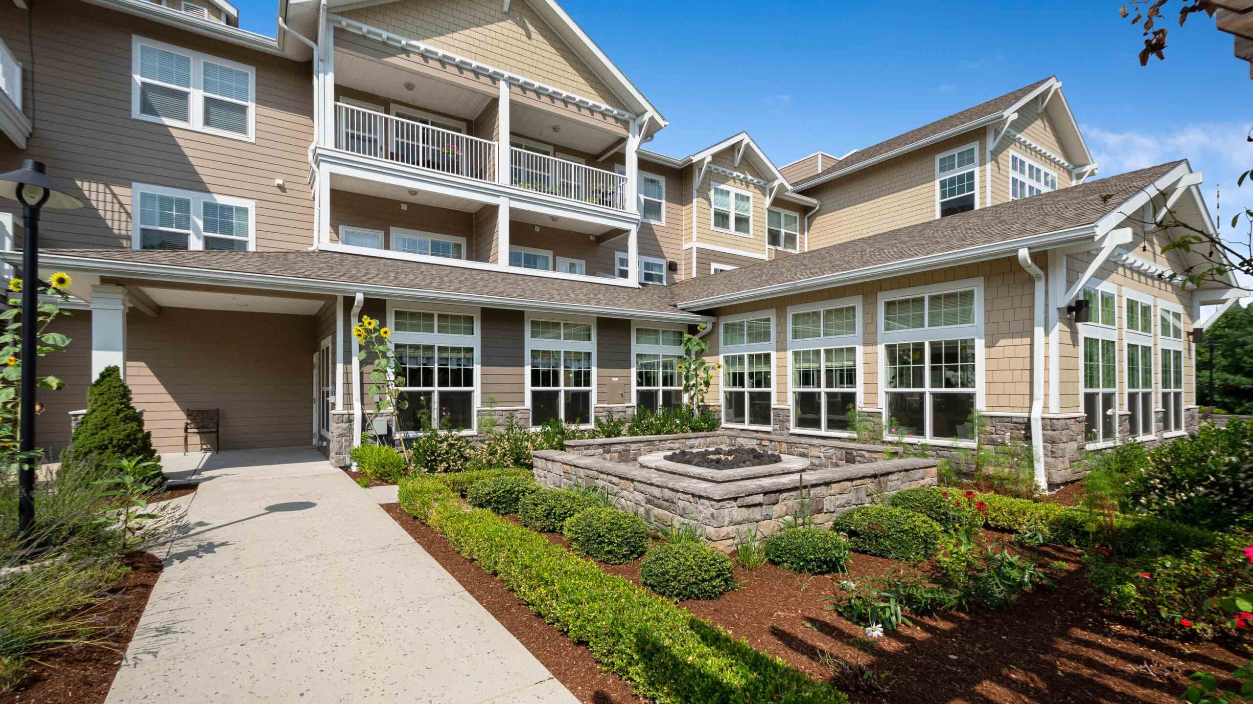 Modern apartment building with beige siding and large windows, featuring a landscaped courtyard with flower beds, green shrubs, and a square stone fountain under a clear blue sky.