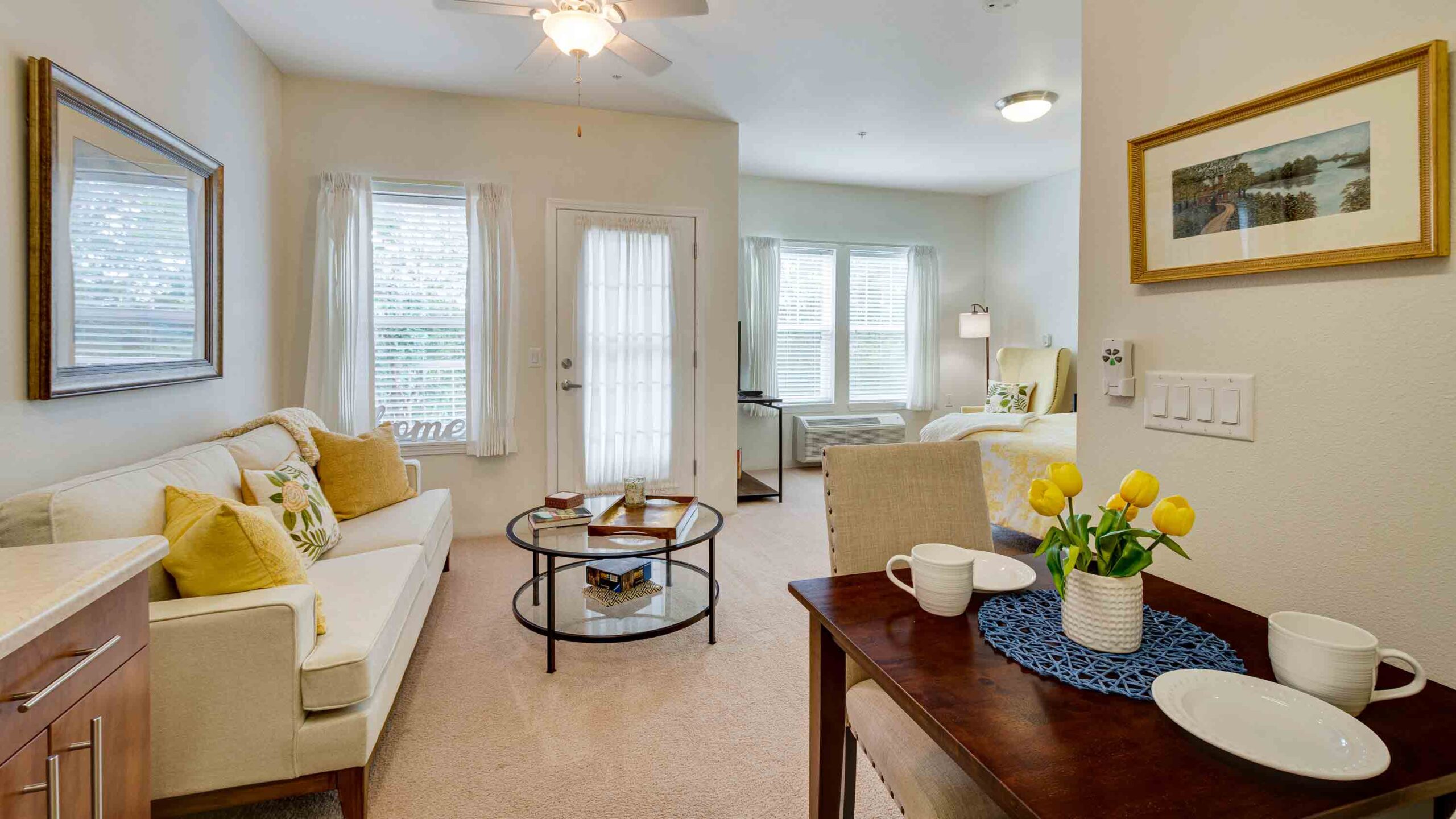 A cozy studio apartment with a sofa, glass coffee table, and bed. A small dining table with two cups and a vase of yellow tulips is in the foreground. Large windows and light decor create a bright, welcoming space.