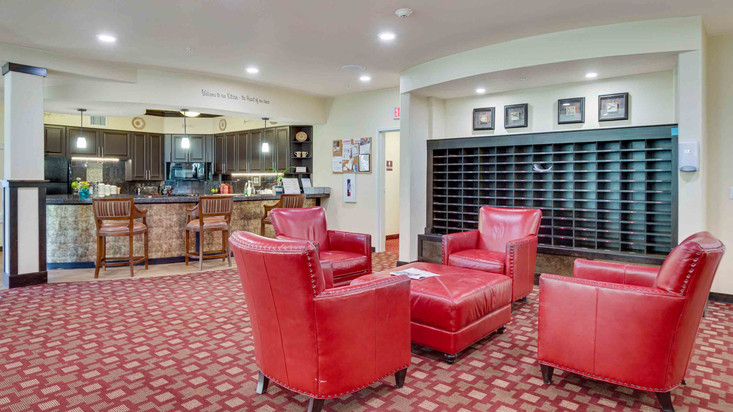 A cozy hotel lobby area with four red leather armchairs around a small table, a patterned carpet, a dark wood reception desk with stools, and mail slots on the wall in the background.