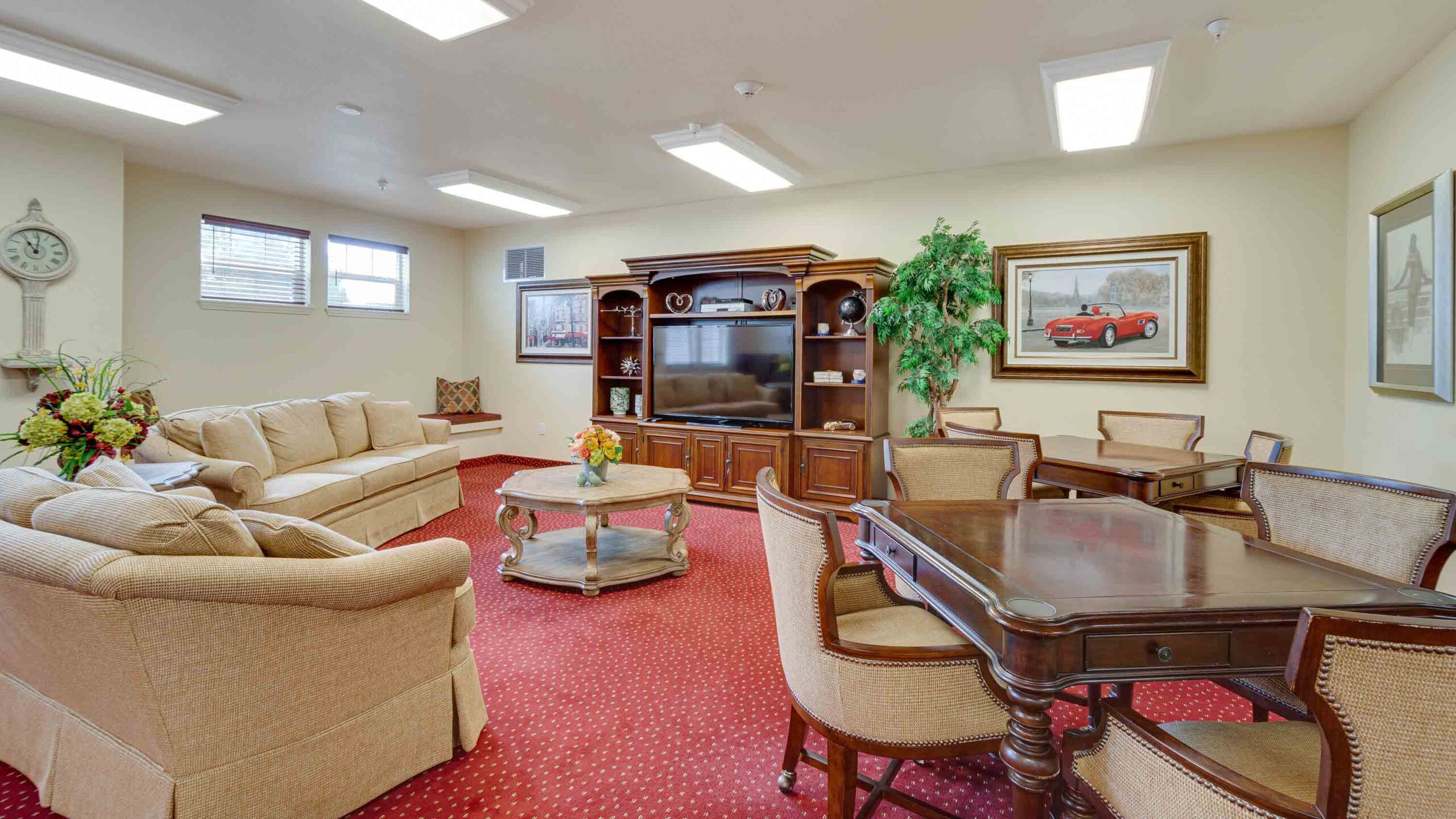 A spacious living room with red carpet, beige sofas, a wooden entertainment center with a TV, a large table with chairs, framed artwork on cream walls, and floral arrangements on tables.