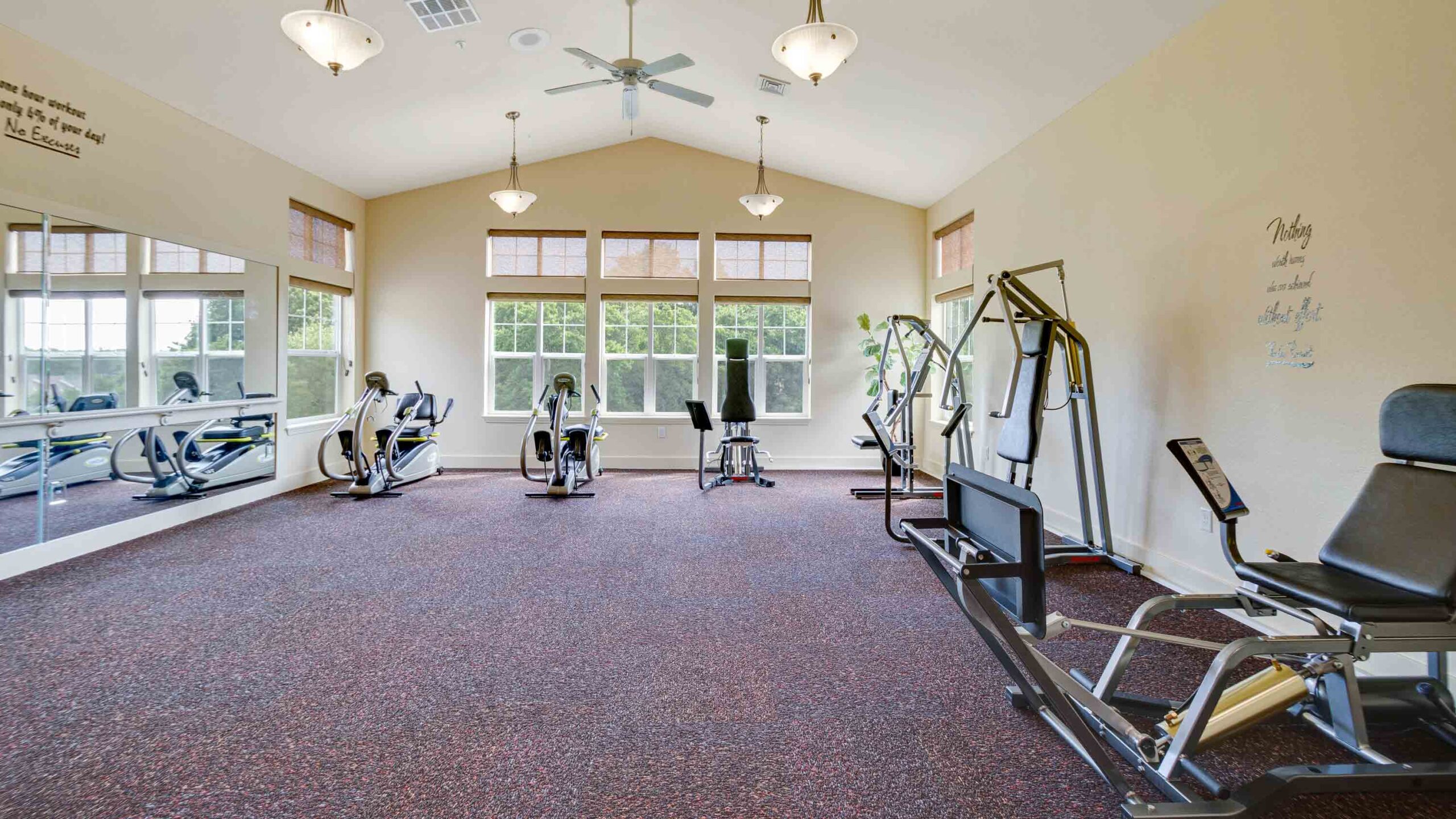 A spacious gym room with large windows, mirrors on one wall, exercise bikes, and strength training machines on a carpeted floor under ceiling lights and fans. Motivational quotes are on the walls.