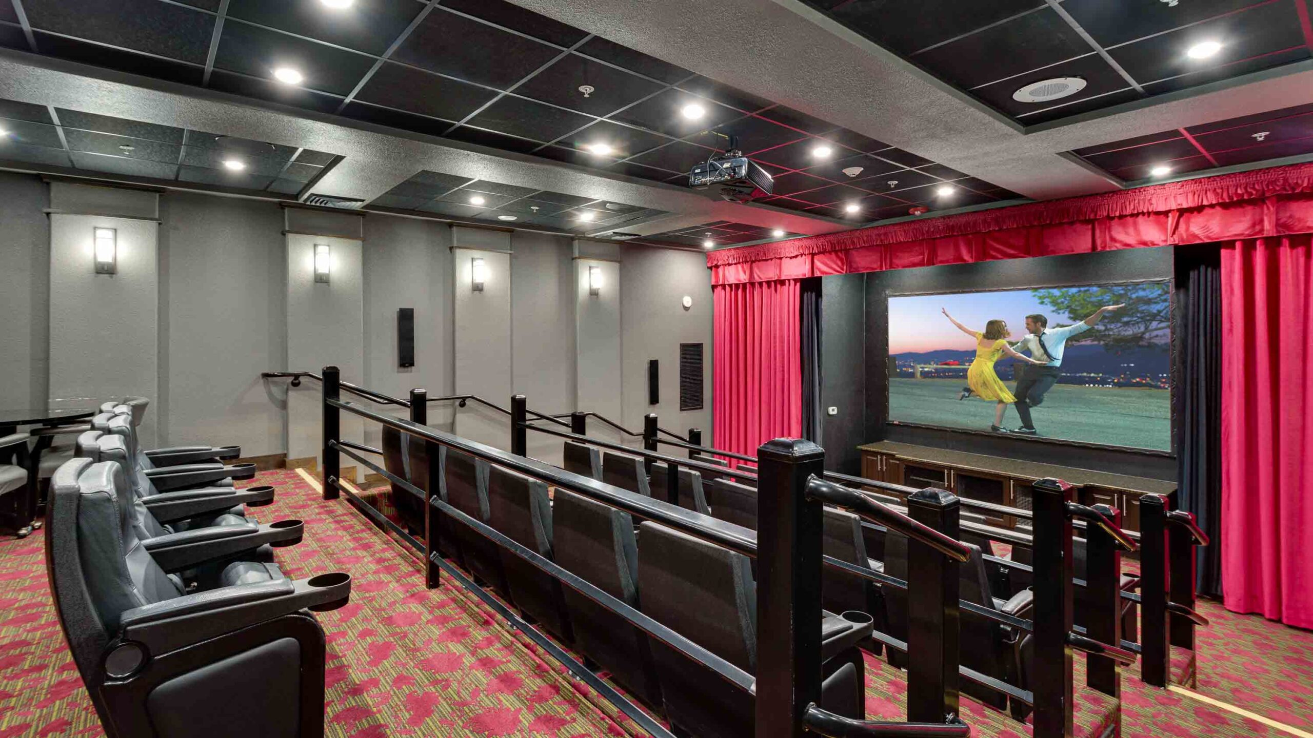 A small modern movie theater with stadium seating, red carpeting, gray walls, and a large screen showing a scene from a musical film. Red curtains frame the screen, and black armchairs fill the rows.