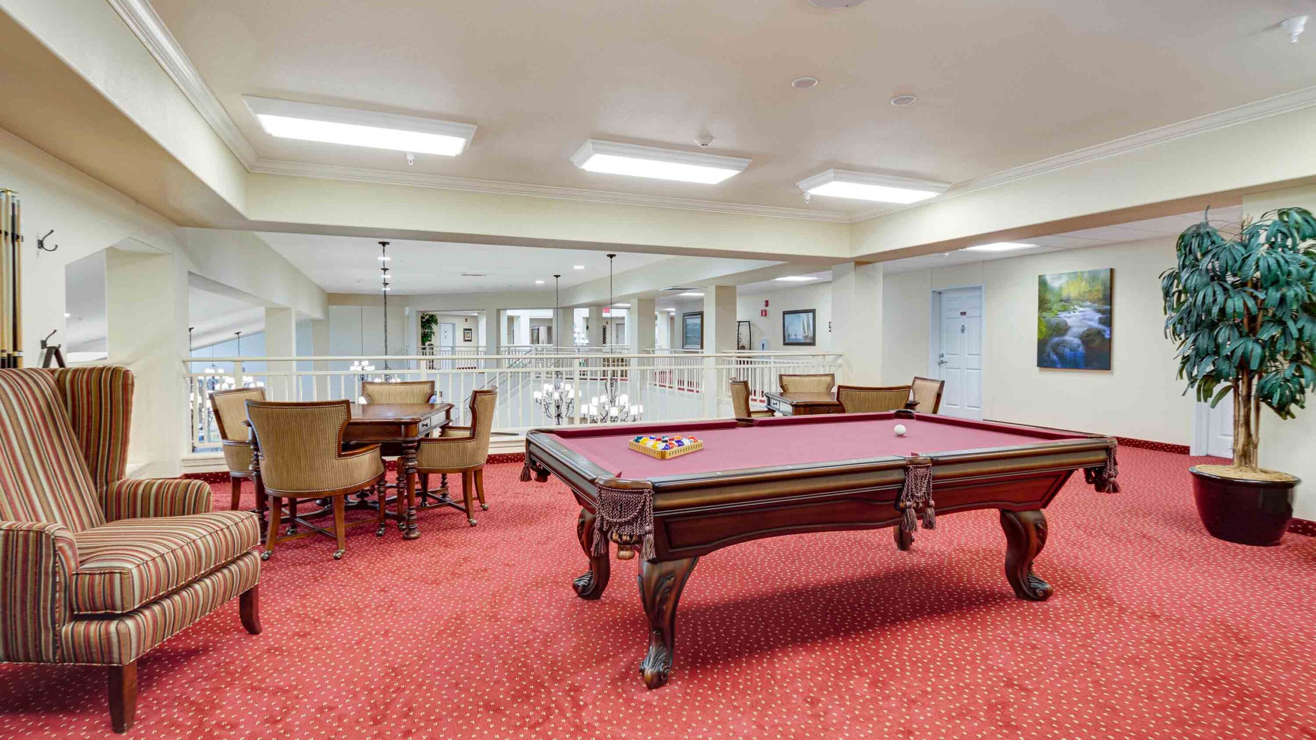 A recreation room with a red-carpeted floor features a pool table set for a game, a striped armchair, a round table with chairs, artwork on the walls, potted plant, and bright ceiling lights.